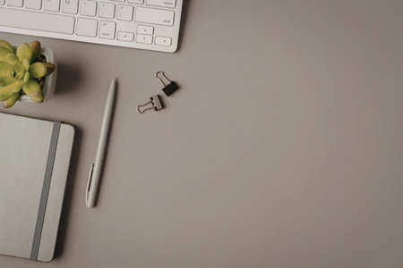 Gray office desk with a keyboard, notebook, pen, binder clips, and a small potted plant creating a...