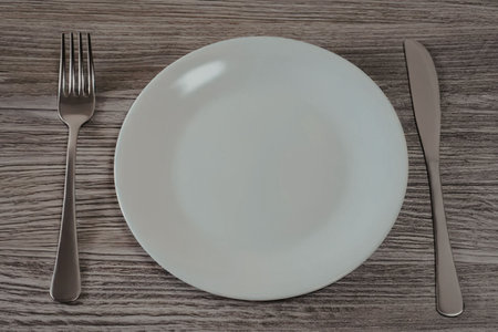 Empty white plate is placed between a fork and knife on a wooden table surface ready for meal...