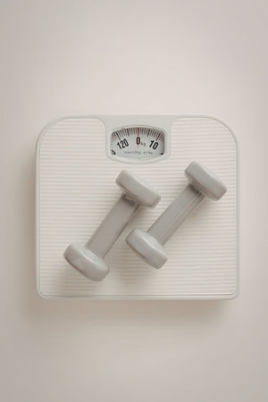 White bathroom scale with two grey dumbbells on it symbolizing fitness and weight management goals...