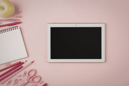 White digital tablet with blank screen on pink background surrounded by stationery including pencils, scissors, paperclips, and green apple, creating a creative workspace atmosphere.の写真素材