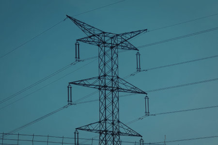 A tall metal power lines tower standing against a bright blue sky with clouds.