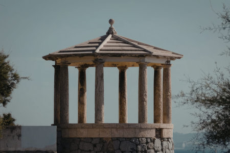 A stone gazebo with columns stands in a park, surrounded by trees and blue sky.