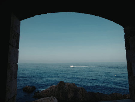 A scenic view of the ocean with boats under a blue sky, seen through a natural rocky arch.