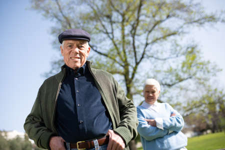Portrait of aged couple posing for camera in park. Happy man in modern clothes and cap smiling looking at camera when woman standing behind him. Relations and leisure activity of aged people conceptの写真素材
