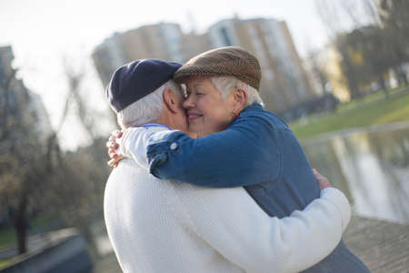 Romantic senior couple resting in spring park. Smiling gray-haired woman in modern jeans clothes and cap hugging mans neck, touching his cheek with her cheek. Relations and love of aged people conceptの写真素材
