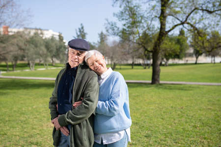 Portrait of aged couple standing on green grass. Happy grey-haired man and woman standing close, woman leaning mans shoulder enjoying warmth of sunny day. Love and active rest of aged people conceptの写真素材