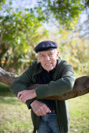 Portrait of aged man under tree in spring park. Smiling Caucasian man with kind eyes standing leaning on tree branch with elbows and looking at camera. Leisure activity of aged people conceptの写真素材