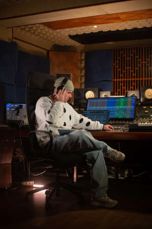 Confident music producer listening to sound track in headphones. Young Caucasian man working at mixing console panel in sound recording studio. Creating music conceptの写真素材