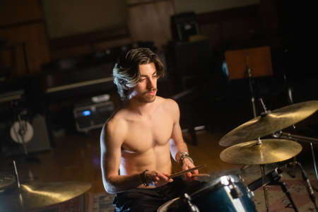 Portrait of young man with moustache playing drum set in garage. Musician sitting shirtless at drum set, training or recording music. Drummer training conceptの写真素材