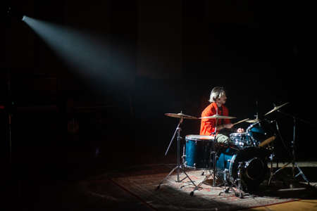 Young man rehearsing on drum set in spotlight in dark studio. Caucasian rock musician recording drums in hall. Drummer training conceptの写真素材