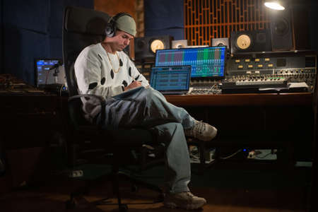 Confident young music producer sitting at mixing console panel and taking notes in notepad. Portrait of Caucasian man with headphones working in sound recording studio. Creating music conceptの写真素材