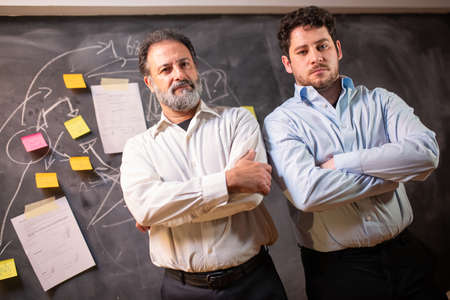 Portrait of attractive colleagues at office. Men in shirts looking at camera, blackboard with writings and paper notes in background. Business, job, late night work conceptの写真素材