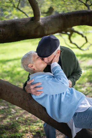 Romantic elderly husband and wife kissing in park. Happy man and woman hugging kissing when woman lying on thick tree branch and enjoying rest. Love, relations and active rest of aged people conceptの写真素材
