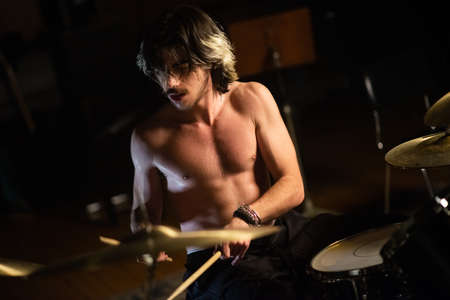 Excited young man sitting shirtless and playing drum set in garage. Rock musician training or recording drums. Drummer training conceptの写真素材