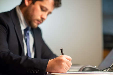 Close-up of blurry businessman writing. Male hand writing with pen on white sheet of paper. Business, job, occupation, late night work conceptの写真素材