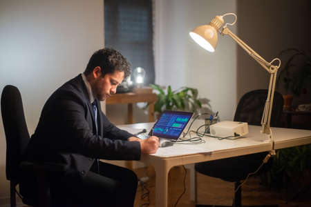 Side view of serious young man working. Man in suit sitting at laptop with charts on screen, writing. Business, job, occupation, late night work conceptの写真素材