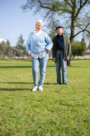 Portrait of modern aged couple posing on field. Attractive woman and man in casual clothes resting in park standing on grass looking aside. Relations and healthy lifestyle of aged people conceptの写真素材