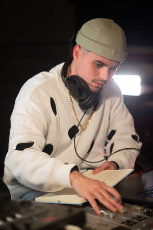 Portrait of concentrated young dj at mixing console panel. Caucasian music producer wearing cap and sweatshirt working in sound recording studio. Creating music, dj conceptの写真素材