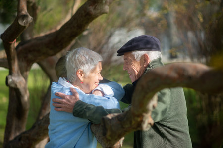 Side view of aged couple resting under trees. Smiling woman looking at man with joy and man hugging her, both standing leaning thick tree branch in park. Love, relations of aged people conceptの写真素材