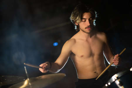 Portrait of concentrated young man playing shirtless on drum set. Caucasian musician training or recording drums in studio. Drummer training conceptの写真素材