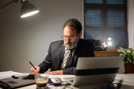 Concentrated bearded man working late at night. Man in suit sitting at table with laptop, thinking....