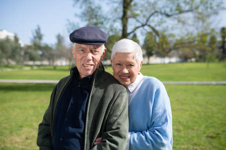 Portrait of senior couple on grassy field. Smiling man in cap and woman resting in park, woman leaning mans shoulder hugging him, both looking at camera. Relations, active rest of aged people conceptの写真素材