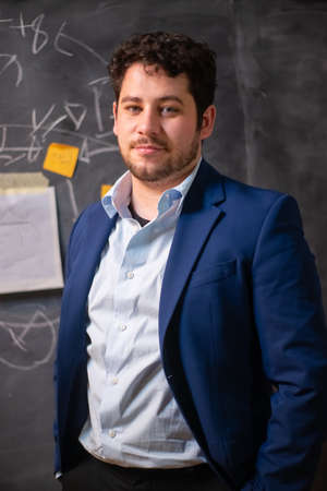Portrait of young handsome businessman at office. Man in suit looking at camera, blackboard with writings and paper notes on it in background. Business, job, late night work conceptの写真素材