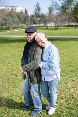 Portrait of aged couple standing on grassy field. Happy man and woman in modern clothes posing together, woman leaning mans shoulder, both looking aside. Love and active rest of aged people conceptの写真素材