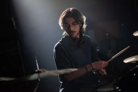 Portrait of concentrated young man playing drum set. Rock musician sitting with headphones recording drums in dark studio. Drummer training conceptの写真素材