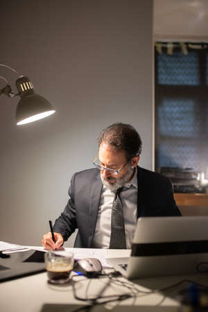 Focused bearded man working late at night. Man in suit sitting at table with laptop, thinking. Business, job, occupation, late night work conceptの写真素材