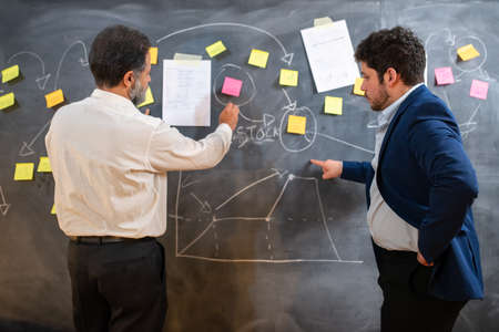 Business partners discussing new project. Two men in suits standing near blackboard with writings...