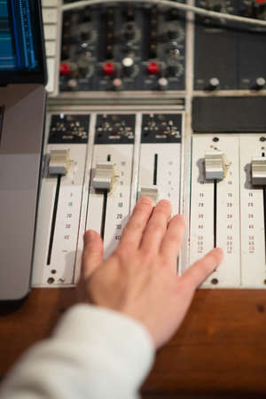 Sound engineer working with mixing console panel in sound recording studio. Close-up of male hand changing volume on mixing desk. Creating music, dj conceptの写真素材