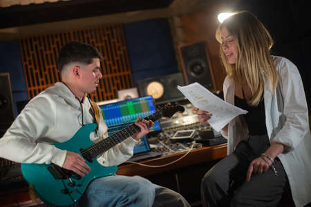 Male guitarist and female singer rehearsing song in studio. Young Caucasian man playing electric guitar and woman singing in sound recording studio. Creating music conceptの写真素材