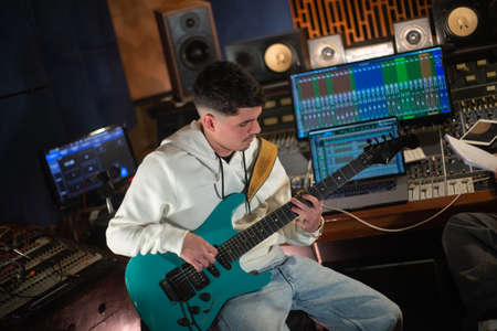Young man sitting at mixing panel and playing electric guitar in sound recording studio. Caucasian guitarist wearing sweatshirt and jeans recording music. Creating music conceptの写真素材