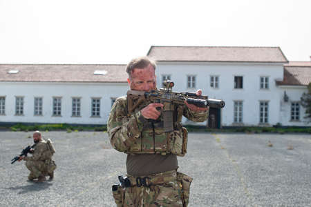 Portrait of bold soldier after battle. Man with blood smears on face standing near house, aiming. Military, army, teamwork, fighting, airsoft conceptの写真素材