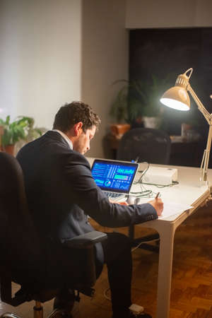 Back view of serious young man working. Man in suit sitting at laptop with charts on screen, writing, working late. Business, job, occupation, late night work conceptの写真素材