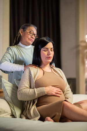 Focused pregnant woman and midwife at home. Woman in casual clothes sitting on bed, Asian doula massaging shoulders. Pregnancy, medicine, home birth conceptの写真素材