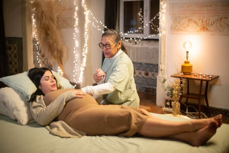 Barefoot pregnant woman and midwife at home. Woman in casual clothes lying on bed, Asian doula...