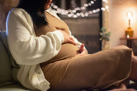 Cropped image of pregnant dark-haired woman sitting on bed. Barefoot woman in dress and cardigan touching big belly. Pregnancy, expectation conceptの写真素材