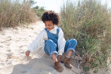 Portrait of African American girl on beach. Female model with curly hair in jeans overall sitting on sand, playing. Portrait, beauty conceptの写真素材