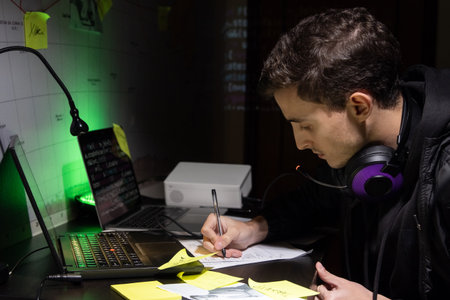 Side view of young man planning hacker attack. Man with headphones sitting at laptop with code,...