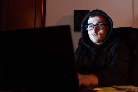 Portrait of young hacker at work. Man in glasses and hood looking at laptop screen, typing. Hacking,...
