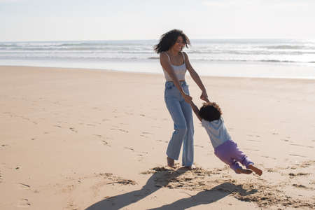 Playful mother and child spending time on beach. African American family walking, twirling around, laughing, fooling around. Leisure, family time, parenthood conceptの写真素材
