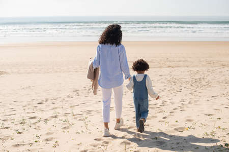Back view of mother and daughter holding hands on beach. African American family spending time together on open air. Leisure, family time, togetherness conceptの写真素材