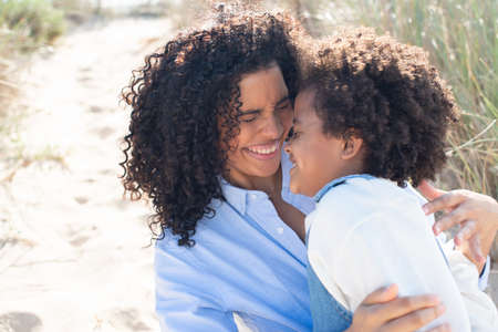 Merry African American mother and daughter on beach. Mother and child in casual clothes sitting on...