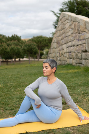 Calm woman with grey hair doing yoga in park. Female model in sportive clothes exercising on mat on...