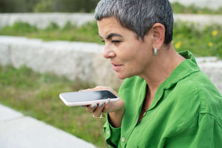 Good-looking senior woman using mobile phone in park. Female model with short grey hair in bright...