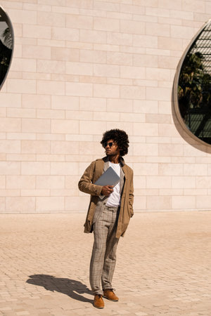 Portrait of smart-dressed African American man outdoors. Man in suit with beard holding laptop, walking. Portrait, city life conceptの写真素材