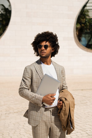 Good-looking African American man outdoors. Man in suit with beard holding laptop, walking. Looking sideways. Portrait, city life conceptの写真素材