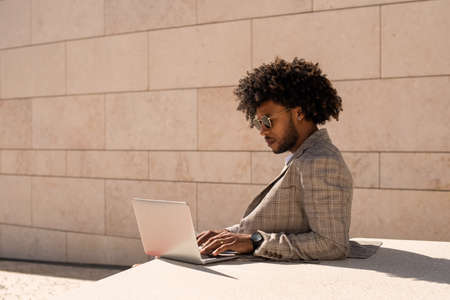 African American man working in open air. Man in suit and sunglasses with beard using laptop. Sitting at terrace or rooftops. Working, manager, technology conceptの写真素材